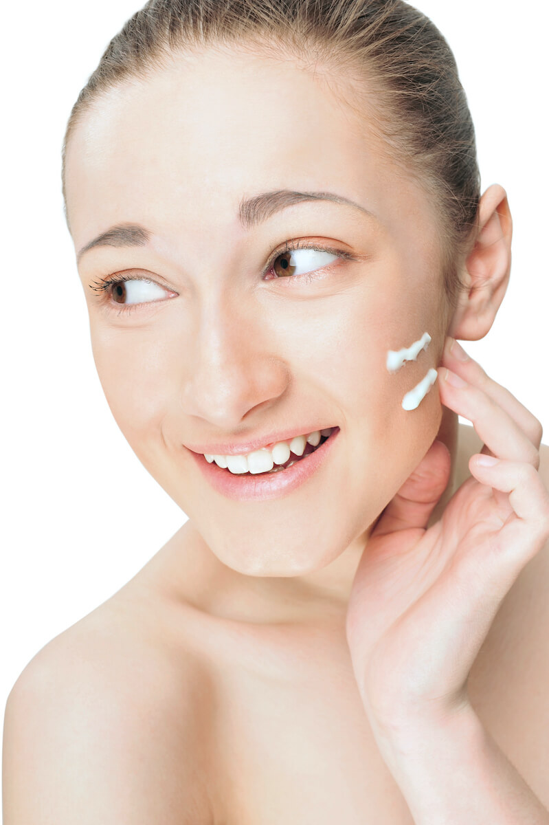 Portrait of a beautiful young woman applying cream to her face and smiling. Isolated white background.